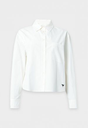 Femme debout, les mains derrière le dos, portant une chemise blanche à boutons, un pantalon beige large et des chaussures noires pointues sur fond blanc.