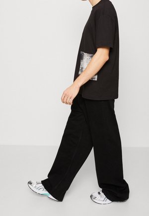 Zwarte katoenen t-shirt met een grafische print op de borst, gecombineerd met losse zwarte broek en witte sneakers met blauwe accenten.