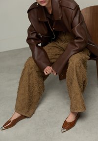 Veste en cuir marron avec épaulettes, pantalon marron shaggy texturé, et chaussures à bout pointu avec embouts métalliques. Décor minimaliste.