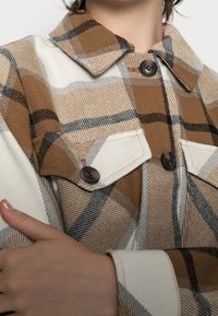 Brown and cream plaid jacket with a collar, featuring large black buttons and dual chest pockets. Soft fabric texture with diagonal lines.