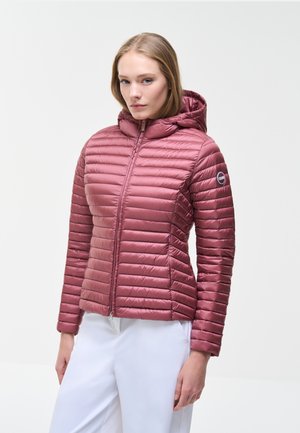 Kvinde iført en mauve quiltet hættejakke med lynlås, stående mod en ensfarvet lys baggrund, med hænderne afslappet ved siderne.