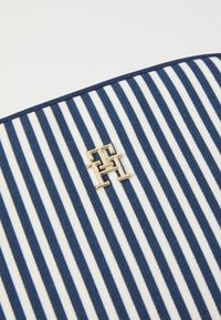 Borsa in tela a righe blu navy e bianco, con un accento in metallo dorato del logo cucito sul davanti. La superficie è liscia e la forma è strutturata.