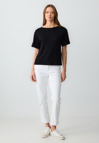Zwart top met korte mouwen en een bootneck ontwerp, gecombineerd met witte broek tot de enkels en witte sneakers, tegen een lichte achtergrond.