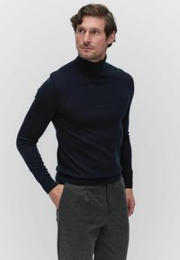 Navy coltrui van zachte gebreide stof, met lange mouwen en een getailleerde pasvorm, gecombineerd met donkergrijze broeken. Geen patronen of versieringen.