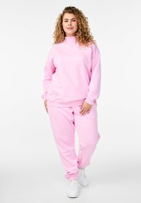 Sudadera rosa y pantalones a juego hechos de una tela suave, con cuello alto y puños acanalados, combinados con zapatillas blancas.