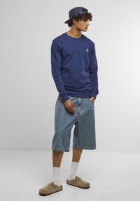 Navy sweatshirt with anchor logo, loose light blue denim shorts, beige clogs, and white socks, styled with a dark blue cap.