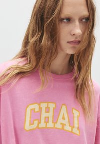 Roze katoenen t-shirt met een relaxte pasvorm, met de tekst "CHAI" in grote gele blokletters op de borst.