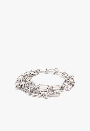 Collier chaîne ton argent avec des maillons allongés, disposé en une boucle lâche sur fond blanc, avec une petite étiquette rectangulaire gravée.