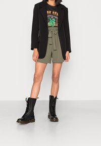 Blazer nero su una t-shirt grafica, pantaloni corti verde oliva con vita regolabile, e stivali neri alti fino al ginocchio con lacci e suole spesse.