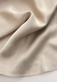 Beige tyg med en mjuk, lätt glänsande yta, som visar mjuka veck och en ren, sydd kant. Lätt och flexibelt material.