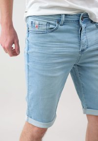 Pantalones cortos de denim azul claro con dobladillo enrollado, diseño de cinco bolsillos y un ligero desgaste. Incluye una etiqueta de logo roja en el bolsillo delantero.
