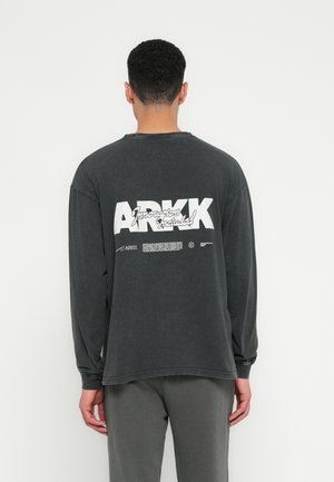 Dugi rukav, izblijedjeli crni majica s velikim bijelim natpisom "ARKK" i grafikom na leđima, s opuštenim krojem i rebrastim manžetama.