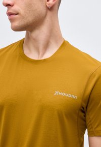 Άνδρας που φοράει μουσταρδί κίτρινη μπλούζα με στρογγυλή λαιμόκοψη και το λογότυπο "Houdini" στο στήθος, κοιτάζει αριστερά σε απλό λευκό φόντο.