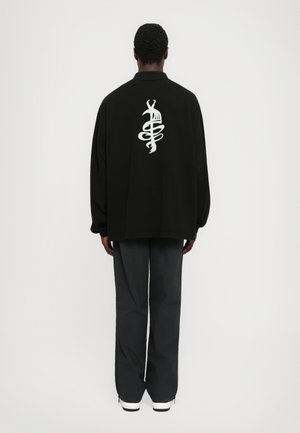 Schwarzer oversized Pullover mit einem weißen Grafikdesign auf dem Rücken. Kombiniert mit dunkelgrauen Hosen und weißen Sneakern.