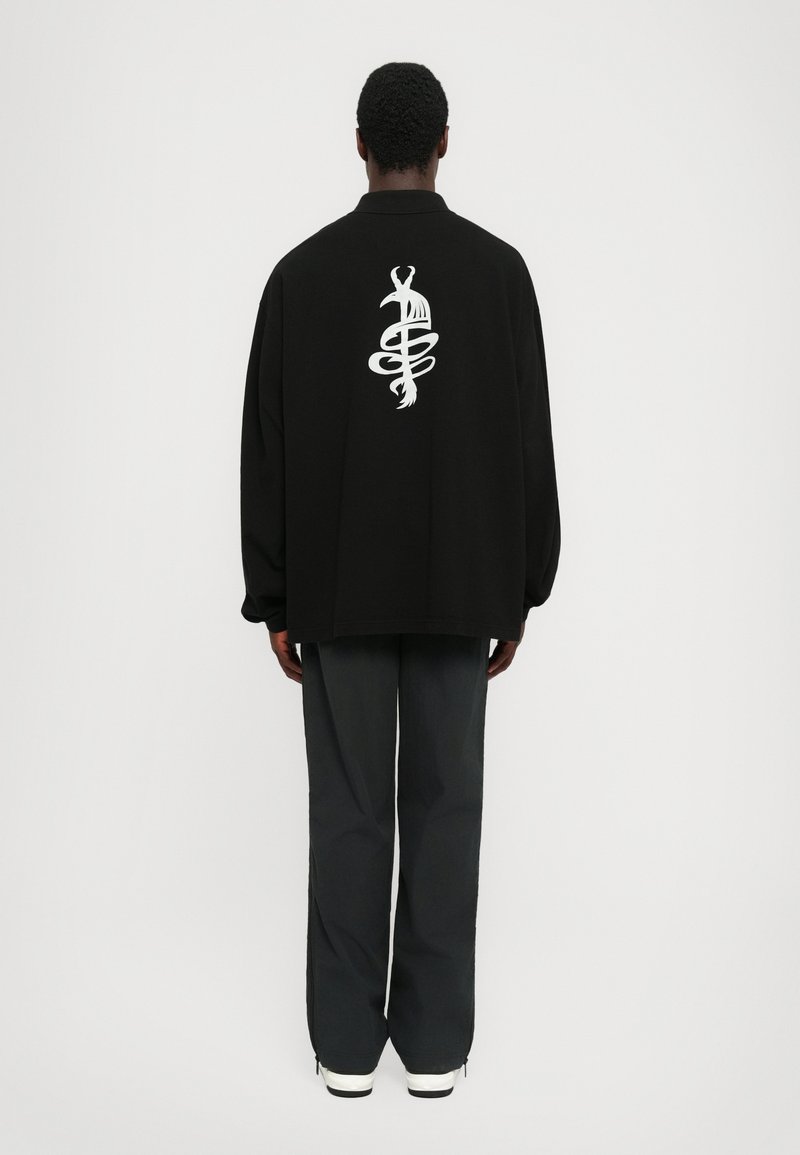 Schwarzer oversized Pullover mit einem weißen Grafikdesign auf dem Rücken. Kombiniert mit dunkelgrauen Hosen und weißen Sneakern.