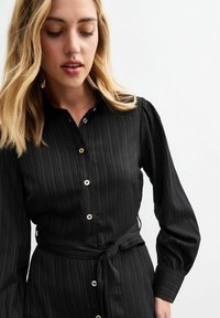 Vestito nero con bottoni, a maniche lunghe a sbuffo e cintura annodata in vita, caratterizzato da righe verticali testurizzate. Presenta bottoni chiari e colletto.