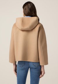 Cappotto di lana beige tagliato con cappuccio, maniche ampie e una texture liscia, abbinato a jeans blu aderenti.