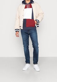 Beige gepolsterte Jacke mit navy- und weißen Streifen an den Bündchen, getragen über einem farblich abgestimmten Hoodie in Rot, Weiß und Navy. Schlanke Jeans aus Denim und weiße Sneakers.