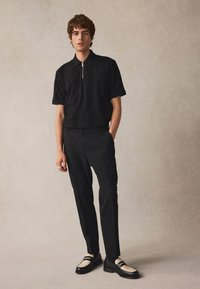 Černé polo tričko s krátkým rukávem a texturovanými vzory, černé kalhoty a dvoubarevné boty kombinující černou a krémovou. Model stojí před neutrálním pozadím.