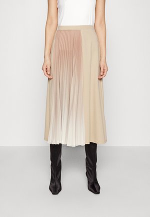 Vrouw met een beige midirok met geplisseerd ombre-paneel en zwarte kniehoge laarzen, staand tegen een effen witte achtergrond.