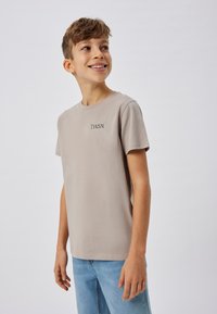 T-shirt en coton beige clair à manches courtes et col rond. Présente un texte brodé noir "T.W.S.N." sur la poitrine. Texture simple et lisse.