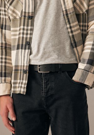 Personne portant une ceinture noire, un jean noir, un t-shirt gris et une chemise beige à carreaux boutonnée, une main dans la poche.