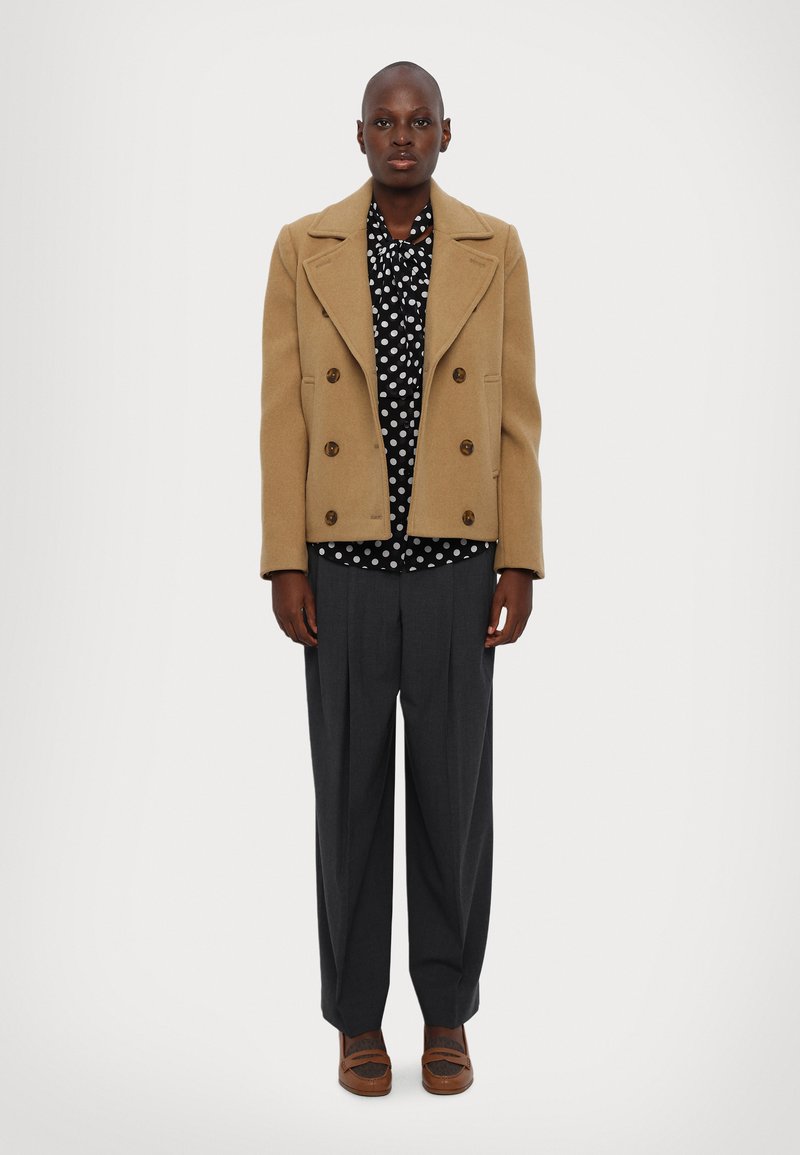 Brauner Woll-Doppelreiher über einem schwarzen Polka-Dot-Hemd, grauen, gefalteten Hosen und braunen Slippern. Vor einem schlichten weißen Hintergrund.