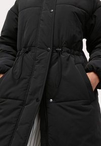 Abrigo acolchado negro con botones a presión, cordones ajustables en la cintura y bolsillos laterales, con una textura de tela suave.