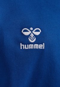 Białe haftowane logo hummel z stylizowaną pszczołą i tekstem na niebieskim tle tkaniny.