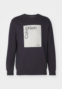 Mörklila sweatshirt med en stor vit rektangulär logotyp med texten "Calvin Klein" och "Est. 1968", tillverkad av mjukt tyg.
