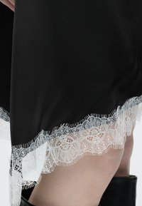 Μαύρη φούστα με λευκή δαντέλα με floral σχέδιο, φορεμένη με μαύρες μπότες, δείχνοντας κοντινό πλάνο του κάτω μέρους του ποδιού και τις λεπτομέρειες του τελειώματος.