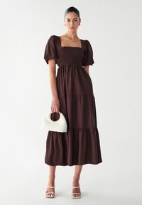 Hnedé maxi šaty s puffovými rukávmi, smúteným korzetom a volánovou sukňou. Spárované s bielou kabelkou v tvare polmesiaca a sandálmi na opätku.