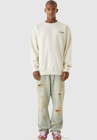 Kremfarget hettegenser med lange ermer og ribbestrikkede mansjetter, kombinert med lyseblå slitte jeans med flere hull. Fotwear inkluderer rosa og hvite sneakers.