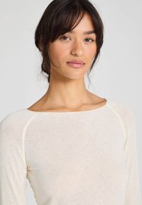 Bluză cu mânecă lungă, cu decolteu tip bardot, fabricată dintr-un material ușor de culoare crem, având modele texturate discrete și cusături contrastante pe margini.