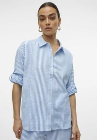 Camicia a maniche lunghe a righe blu e bianche con maniche arrotolate e colletto. Realizzata in tessuto leggero con una vestibilità comoda.