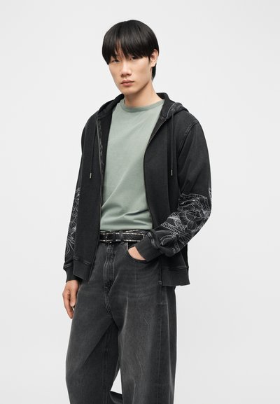 Νέος άνδρας που φοράει μαύρο φούτερ με κουκούλα και σχέδια με λουλούδια στα μανίκια, ανοιχτοπράσινο μπλουζάκι, μαύρη ζώνη με τρουκς και φαρδιά σκούρα γκρι τζιν.