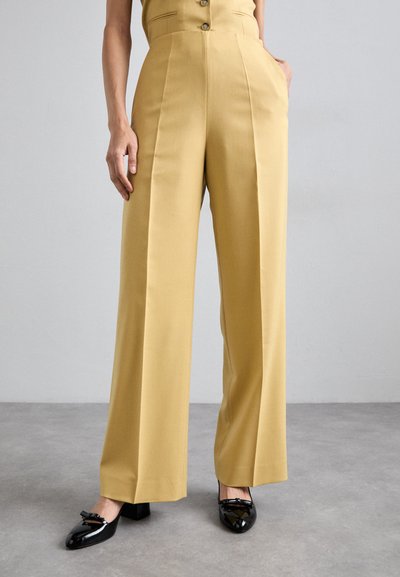 Wide-leg trousers in mustard yellow fabric with sharp creases and a structured waist. Paired with black patent leather shoes.