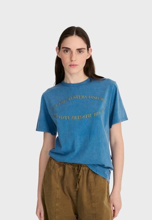 Jeune femme aux longs cheveux foncés portant un t-shirt bleu imprimé de texte et un pantalon marron à cordon, sur fond gris clair.