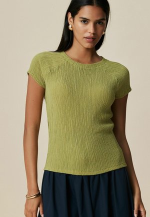 Short-sleeve green top with textured fabric, featuring a round neckline and fitted silhouette. Worn with a navy skirt.
