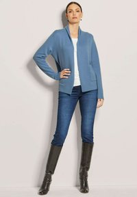 Hellblauer Cardigan mit Schalkragen und gerippten Bündchen, kombiniert mit dunkelblauen Skinny-Jeans und kniehohen schwarzen Stiefeln.