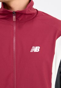 Mørkerød sportjakke med høj krave, lynlås-lukning og kontrastfarver i sort og hvid. Har et hvidt New Balance-logo.