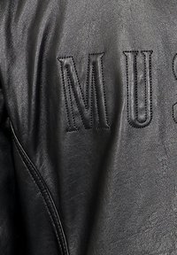 Zwarte leren jas met geborduurde letters "MUS" in een contrasterende steek, met een gladde textuur en een getailleerde pasvorm.