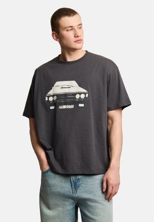 Młody mężczyzna z krótkimi włosami, ubrany w ciemnoszarą koszulkę z nadrukiem przedniej części zabytkowego samochodu oraz jasnoniebieskie dżinsy, stoi z jedną ręką w kieszeni.