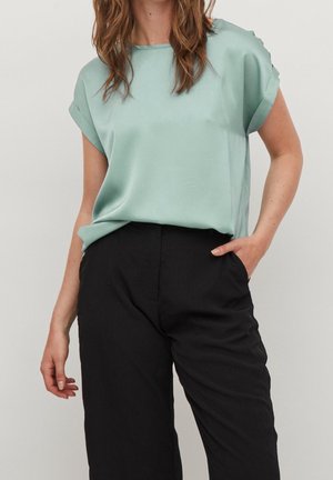 Jasnozielona bluzka z krótkim rękawem w gładkim materiale o luźnym kroju, zestawiona z spodniami o wysokim stanie w ciemnym kolorze. Rękawy lekko podwinięte.