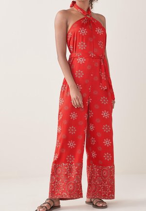 Femme portant une combinaison rouge à col halter avec des motifs floraux blancs et des jambes larges, associée à des sandales plates marron foncé.