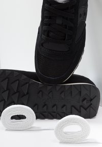 Baskets noires avec semelle texturée reposant sur le côté, accompagnées de deux lacets blancs soigneusement enroulés sur une surface blanche.