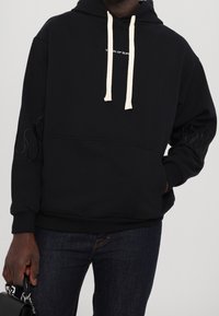 Persona que lleva una sudadera negra con cordones blancos, detalles de llamas en las mangas, jeans oscuros y sostiene un pequeño bolso negro.