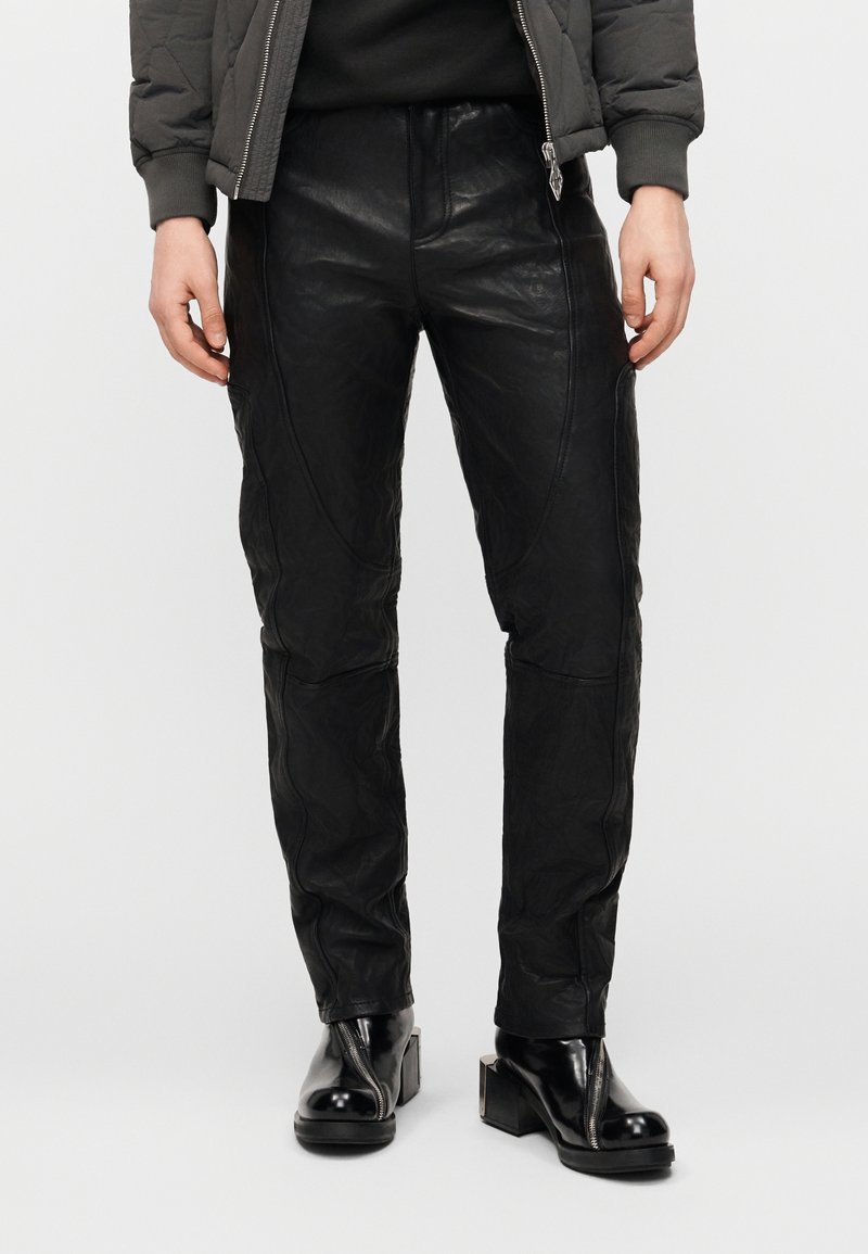 Άτομο που φοράει μαύρο δερμάτινο παντελόνι, μαύρα μπότες με τακούνι και φερμουάρ μπροστά, και σκούρο καπιτονέ μπουφάν με λεπτομέρεια φερμουάρ.