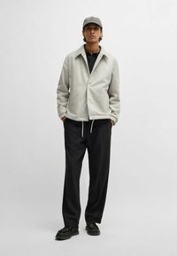 Giacca in misto lana grigio chiaro con vestibilità rilassata e bottoni frontali, abbinata a pantaloni neri e scarpe scure. Un berretto semplice completa l'outfit.