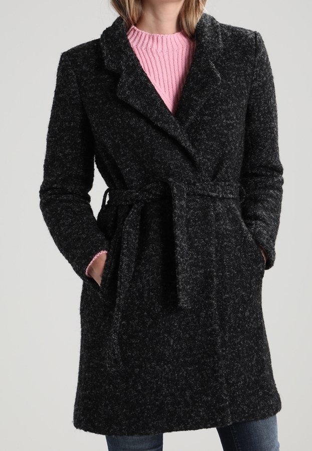 Femme portant un manteau texturé gris foncé ceinturé par-dessus un pull côtelé rose et un jean bleu, les mains dans les poches du manteau.